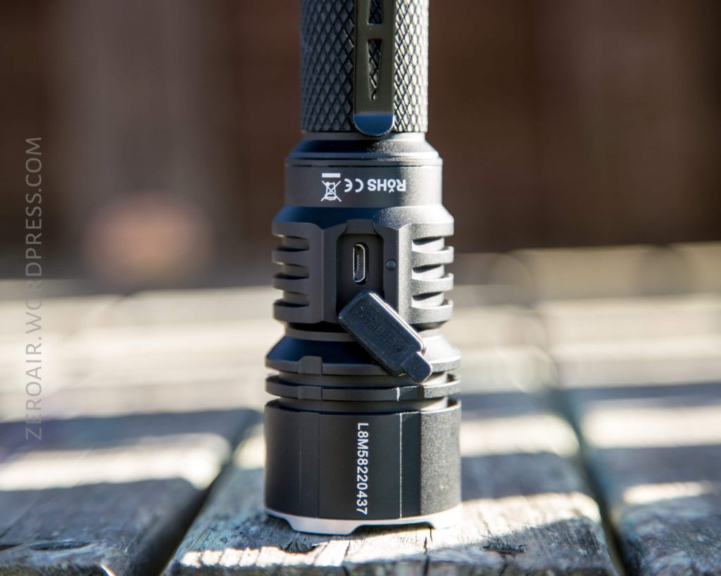 A close-up of an Acebeam L16 Flashlight standing upright on a wooden surface, showcasing its textured grip, USB charging port with cover, and serial number L8MB5024037 on the lower part.