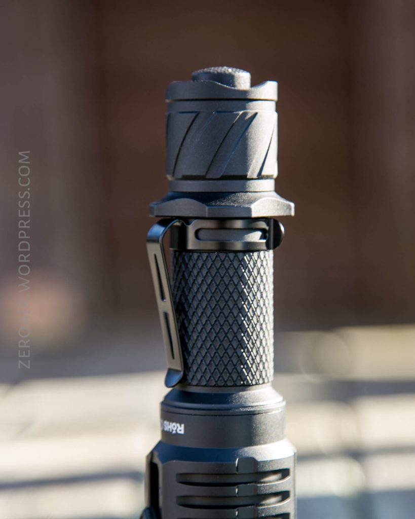 A close-up view of the Acebeam L16 Flashlight standing upright outdoors, with sharp focus on its knurled grip and pocket clip; the background is blurred.