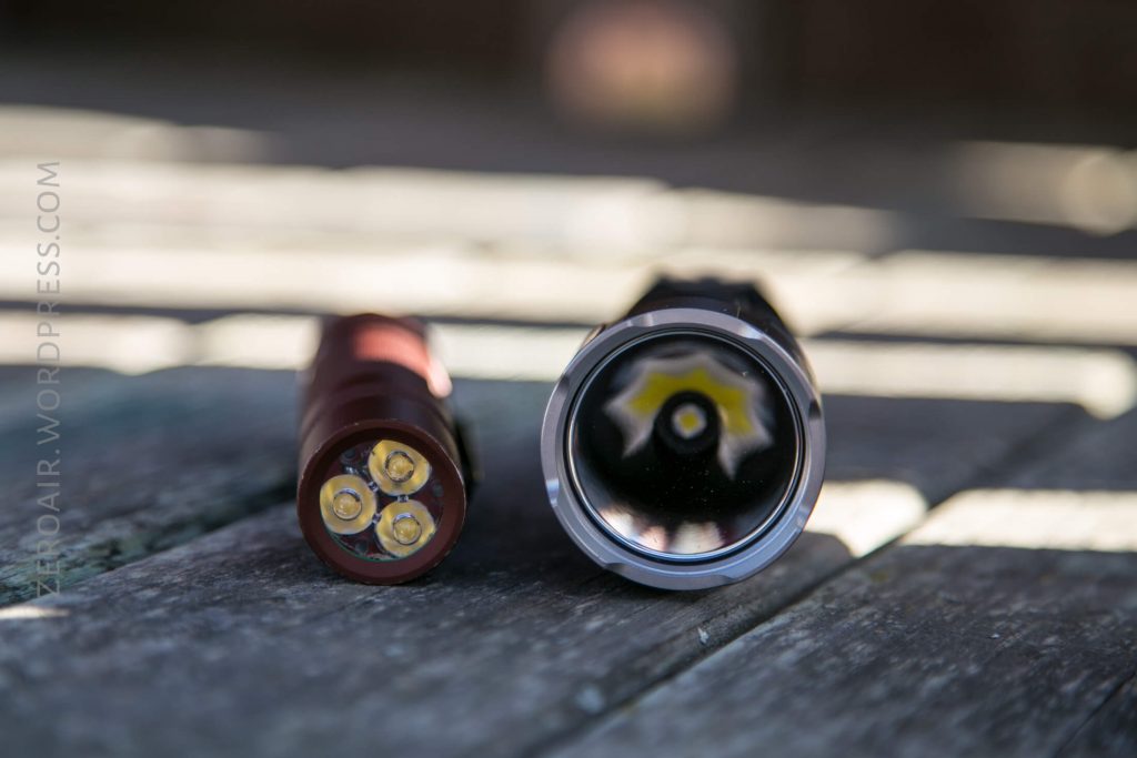 Two disassembled parts of an Acebeam L16 Flashlight rest on a wooden surface in sunlight; the left part displays three exposed gold battery contacts, while the right reveals the reflector and LED inside the flashlight head.