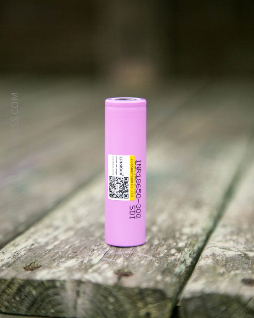 A single pink cylindrical lithium-ion battery labeled INR18650-30Q stands upright on a weathered wooden surface. The battery has a white barcode label and black text printed along its side.