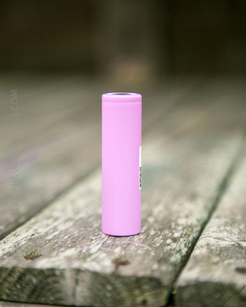 A single pink cylindrical battery standing upright on a weathered wooden surface with a blurred background.