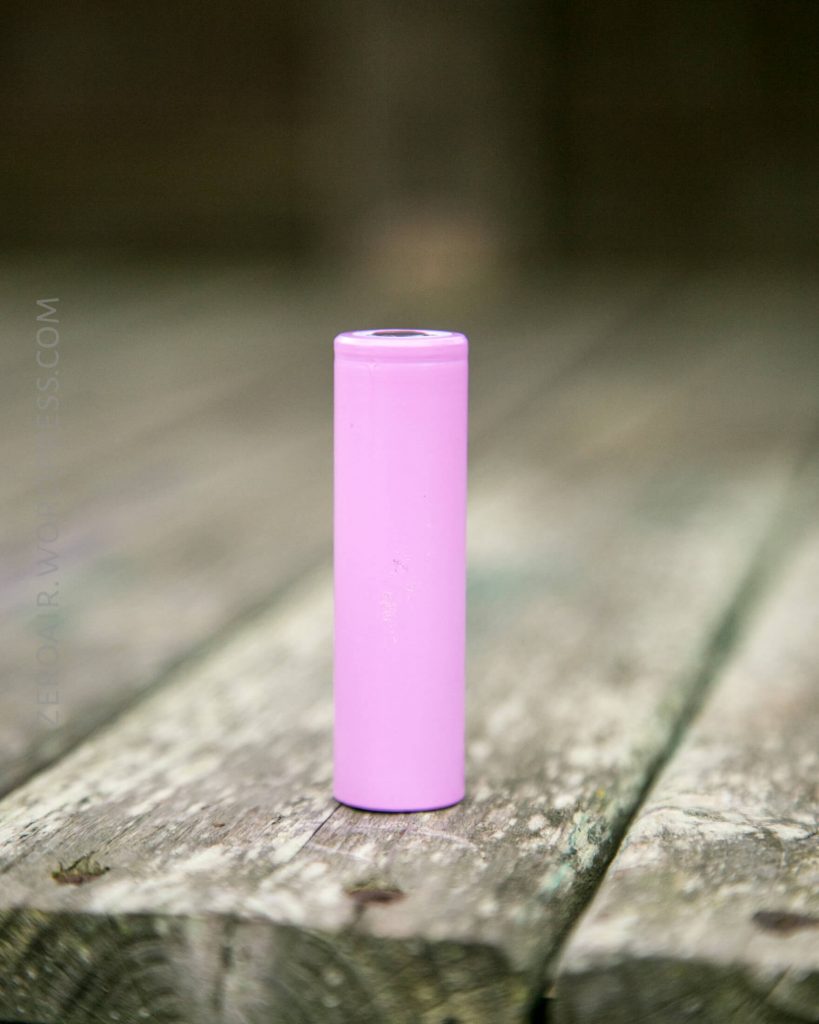 A single pink cylindrical battery standing upright on a weathered wooden surface with a blurred background.