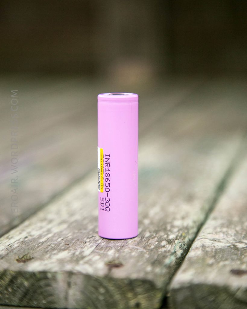 A single pink cylindrical battery standing upright on a weathered wooden surface with a blurred background. The battery has some text printed on its side.