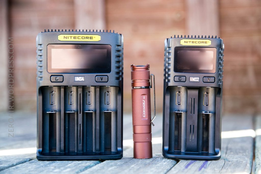 Two black Nitecore battery chargers of different sizes are placed on a wooden surface outdoors, with a brown Fenix flashlight standing upright between them. A blurred wooden fence is in the background.