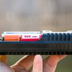 A close-up of a persons hand holding a black battery charger with two cylindrical batteries inside, one red and one orange, against a blurred outdoor background.