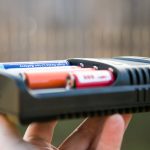 A close-up view of a hand holding a Nitecore battery charger with two cylindrical batteries, one blue and one orange, inserted in the device against a blurred background.