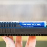A hand holds a black battery charger with a blue cylindrical 21700 lithium-ion battery installed. The battery label reads 3750mAh, 3.7V, 13.88Wh. The background is blurred.