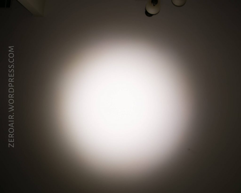 A bright white spotlight shines on a blank wall in a dark room. “ZEROAIR.WORDPRESS.COM” is written vertically on the left side of the image. Two ceiling light fixtures are partially visible at the top right.