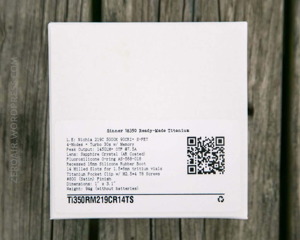 A white box with a detailed label listing technical specifications, QR codes, and a product code. The label includes information about materials, battery, screws, and clip, with a blurred wooden surface in the background.