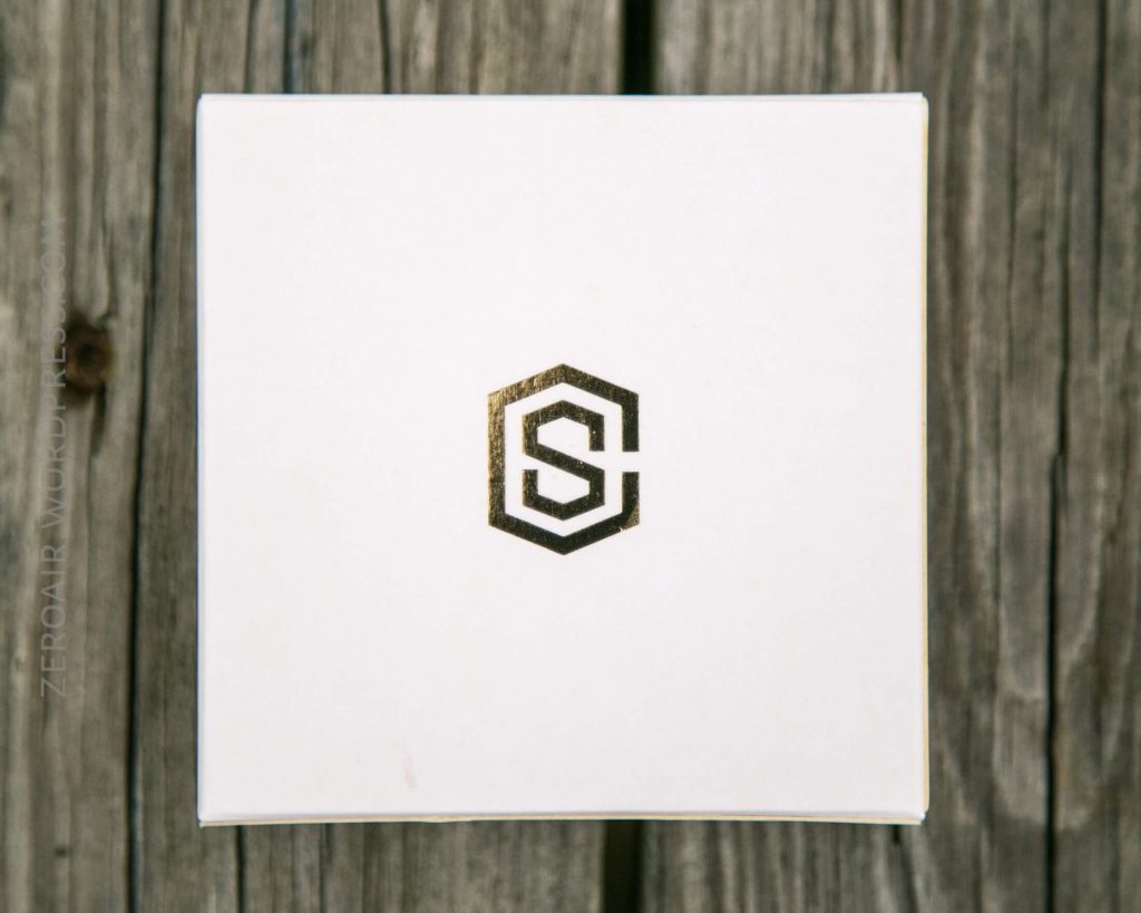 A white square box with a metallic gold geometric logo resembling the letters “S” and “C” intertwined, placed on a weathered wooden surface.