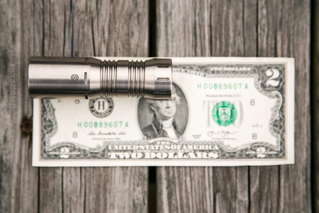 A metallic flashlight is placed horizontally on top of a two-dollar bill, which lies on a weathered wooden surface.