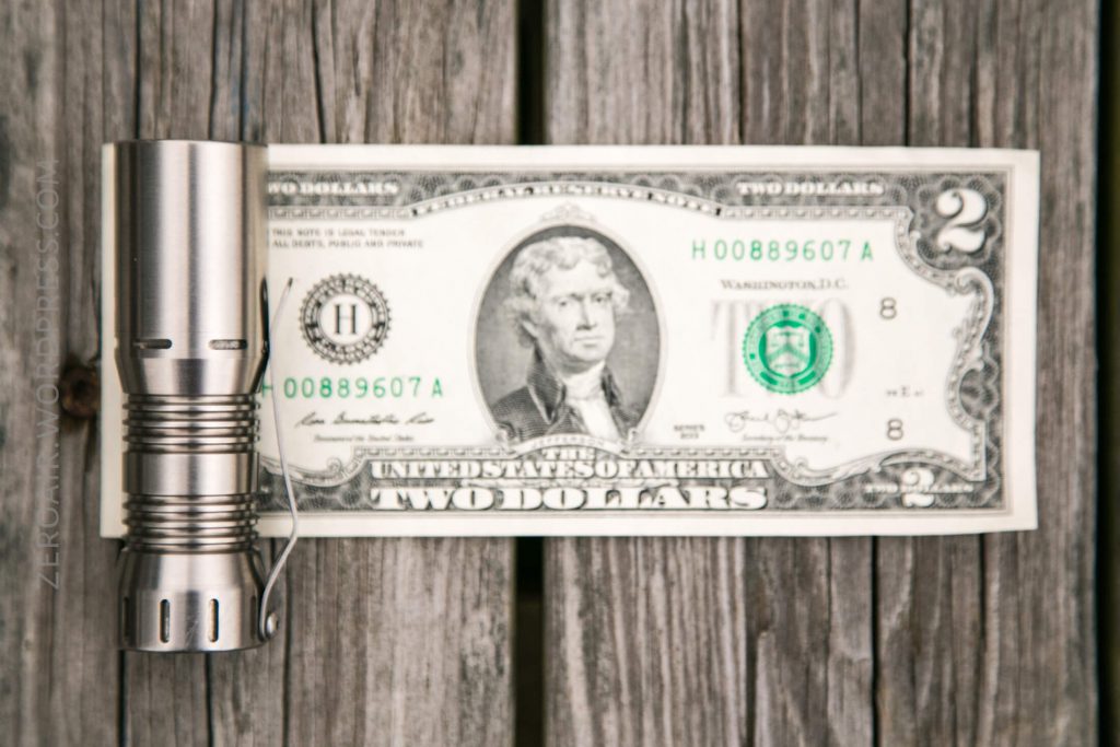 A metal flashlight lies next to a two dollar bill on a weathered wooden surface, with the flashlight aligned along the left edge of the bill.