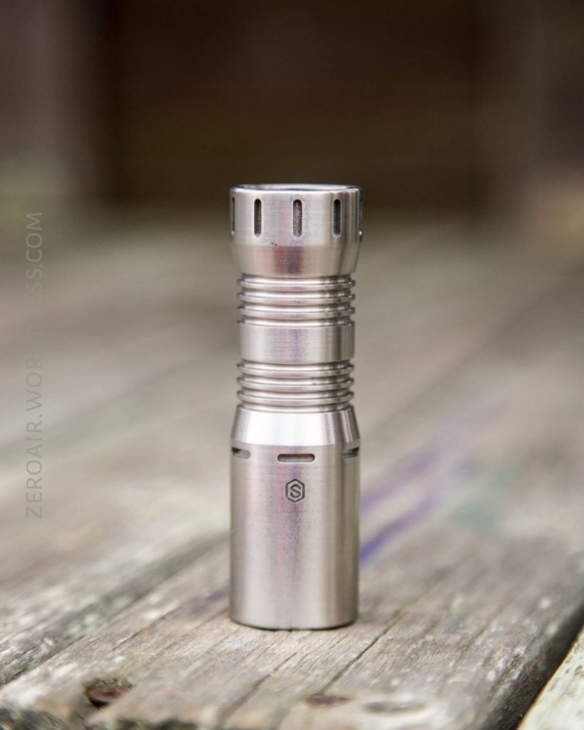 A stainless steel flashlight stands upright on a weathered wooden surface, with the brand logo visible on its side. The background is blurred and the focus is on the flashlight.