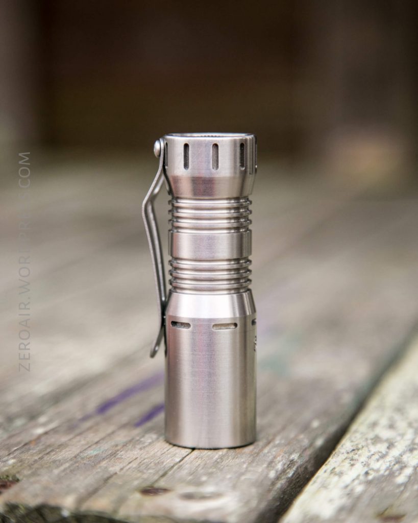 A small, metallic flashlight with a pocket clip standing upright on a weathered wooden surface. The background is blurred.