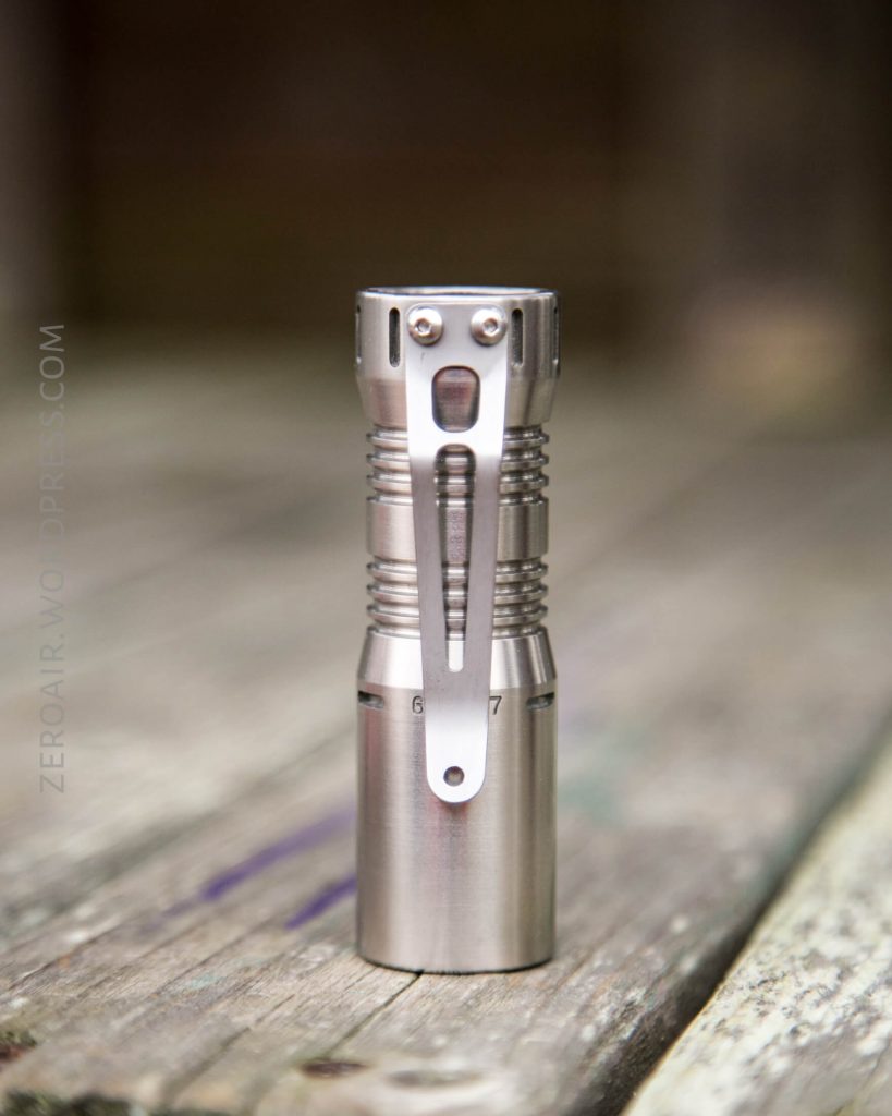 A stainless steel flashlight with a metal pocket clip stands upright on a weathered wooden surface. The background is blurred.