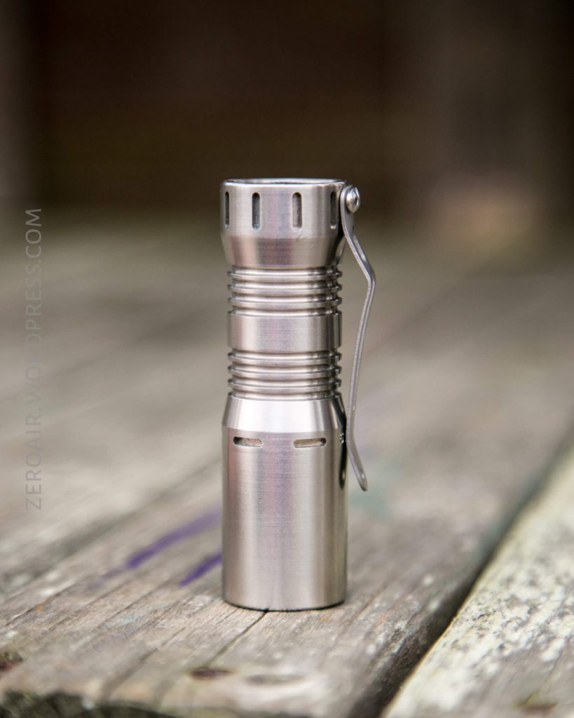 A small, metallic flashlight with a pocket clip stands upright on a weathered wooden surface. The background is blurred, drawing focus to the flashlight.
