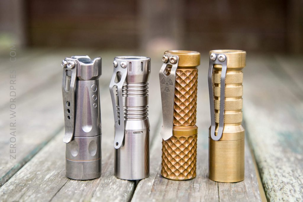 Four metal flashlights, each with a side pocket clip, stand upright in a row on a weathered wooden surface. The flashlights vary in finish and texture, including smooth, brushed, and knurled patterns.