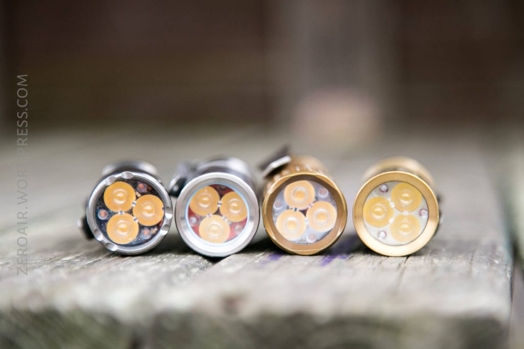 Four small, cylindrical flashlights are positioned upright on a weathered wooden surface, each with three LED emitters visible in their round front faces. The bodies are silver, gray, gold, and brass in color.