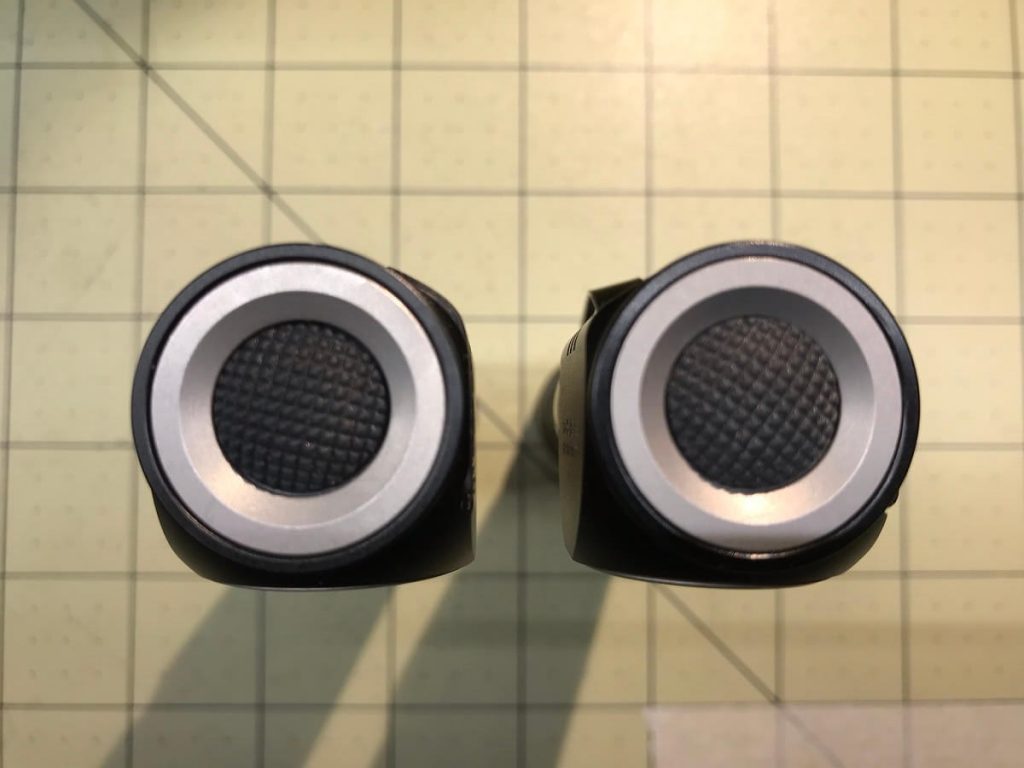 Two black cylindrical objects with textured circular surfaces and metallic outer rings are shown side by side on a grid-patterned background.