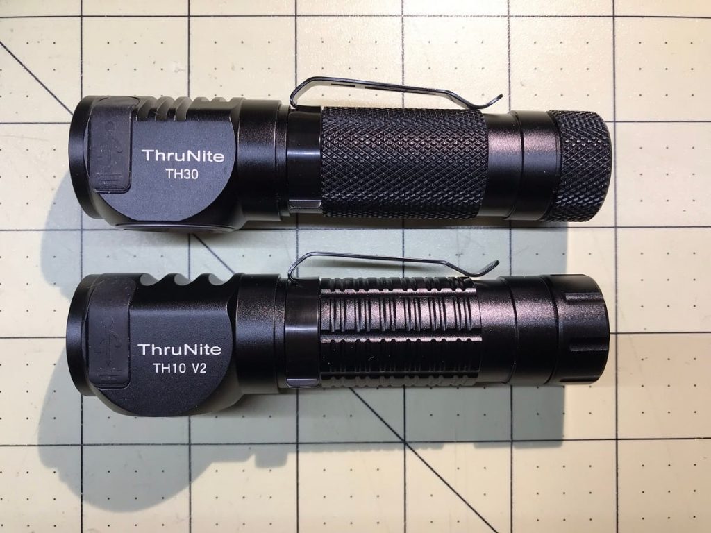 Two black ThruNite flashlights, models TH30 and TH10 V2, are placed side by side on a grid-patterned surface. Both have pocket clips and textured grips, with labels visible near the heads of the lights.