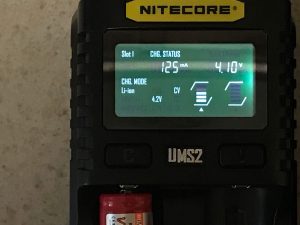 Close-up of a Nitecore UMS2 battery charger display showing charging status: 125 mA current, 4.10 V voltage, charging mode Li-ion 4.2V CV. Part of a red battery is visible in the charger slot below the screen.