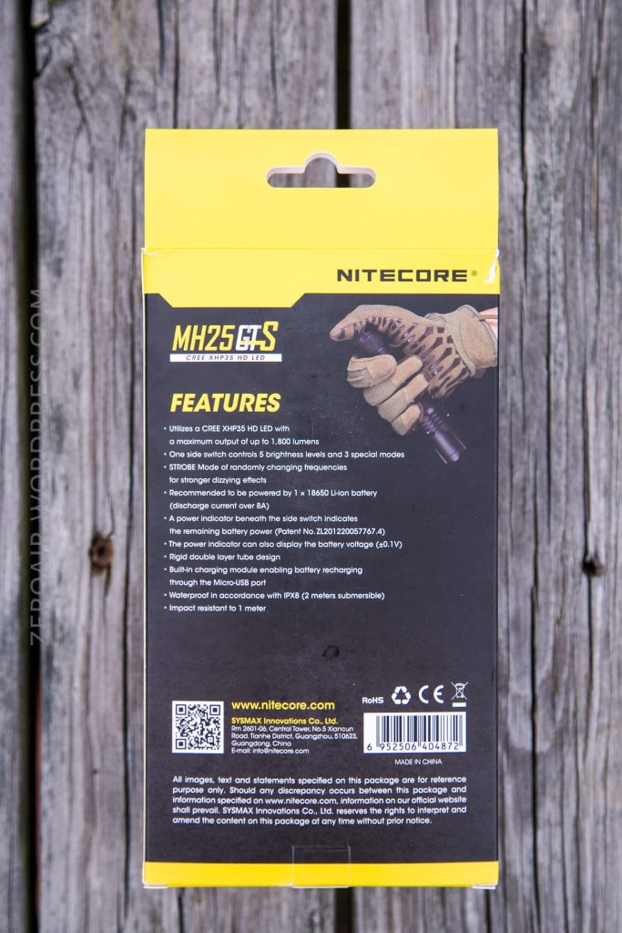 A close-up of the back of a Nitecore MH25GTS flashlight box on a wooden surface, listing features and manufacturer details, with an image of a gloved hand holding the flashlight.