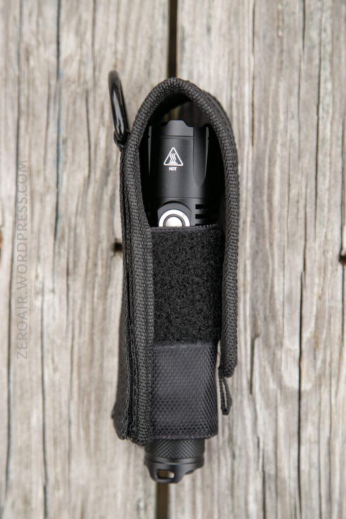 A black flashlight is secured inside a black fabric holster with a Velcro flap, resting vertically on weathered wooden boards. The flashlights power button and warning symbol are visible.