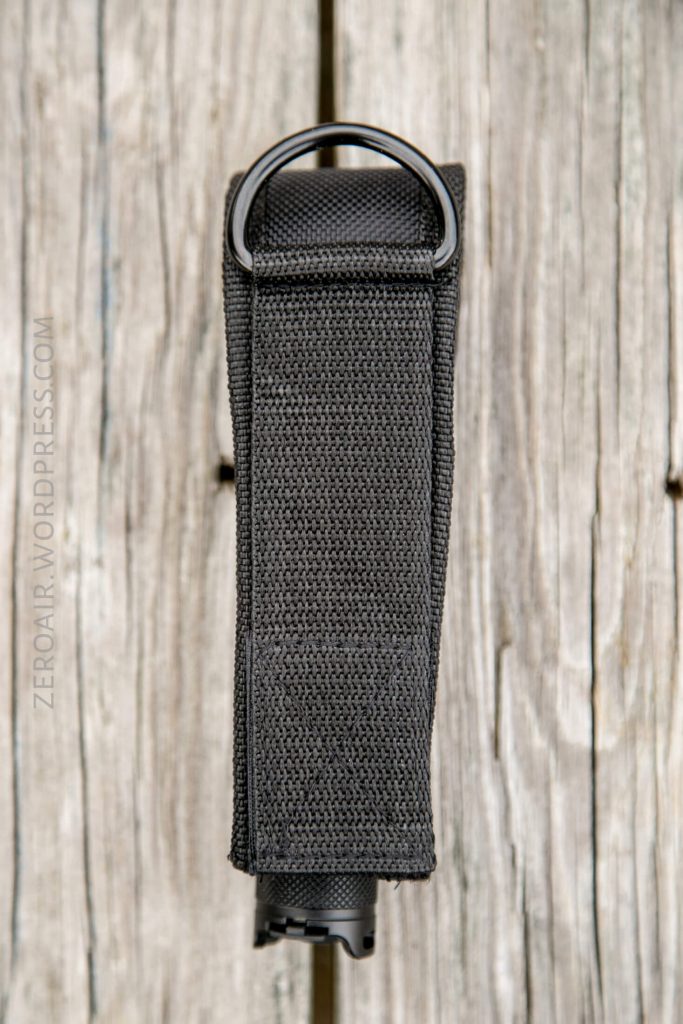A black, textured nylon pouch with a D-ring at the top, placed vertically on a weathered wooden surface. The pouch is closed and stitched with visible threading.