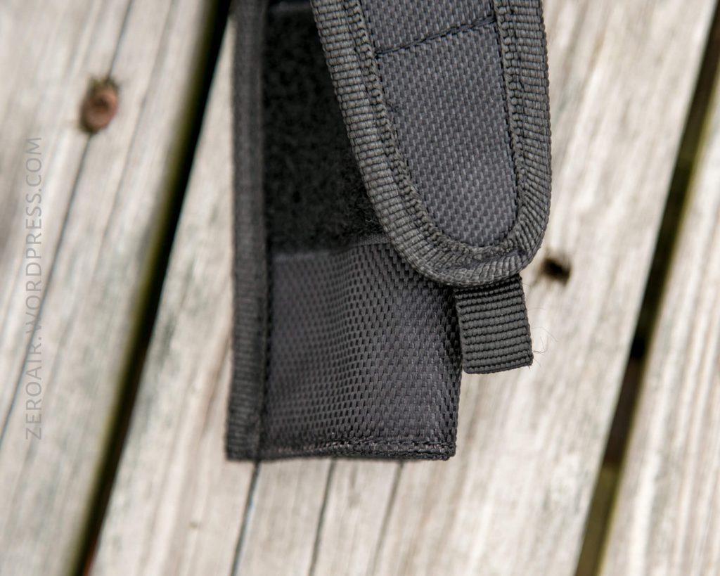 A close-up of a black textured nylon pouch with a flap, resting on weathered wooden boards. The pouch has reinforced stitching and a small fabric loop. The words ZEROAIR.WORDPRESS.COM are visible along the left edge.