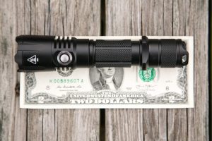 A black tactical flashlight is placed horizontally on top of a two-dollar bill, which rests on a wooden surface. The flashlight is slightly longer than the bill’s width.