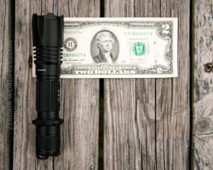 A black tactical flashlight is placed next to a two-dollar bill on a weathered wooden surface. The flashlight is roughly the same length as the bill. The words ZEROAIR.WORDPRESS.COM appear on the left.