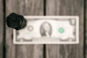 A small black flashlight stands upright on a blurred one-dollar bill, placed on a wooden surface. The website ZEROAIR.WORDPRESS.COM is visible along the left edge of the image.