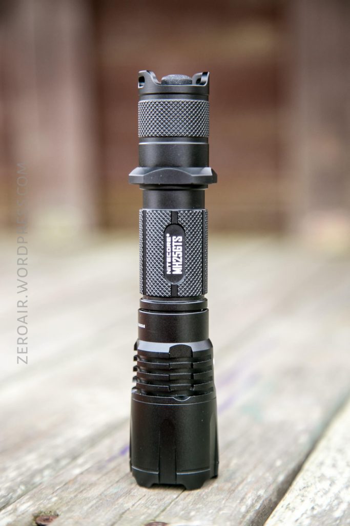 A black tactical flashlight stands upright on a wooden surface, with textured grip sections and the label WUBEN visible on the body. The background is blurred.