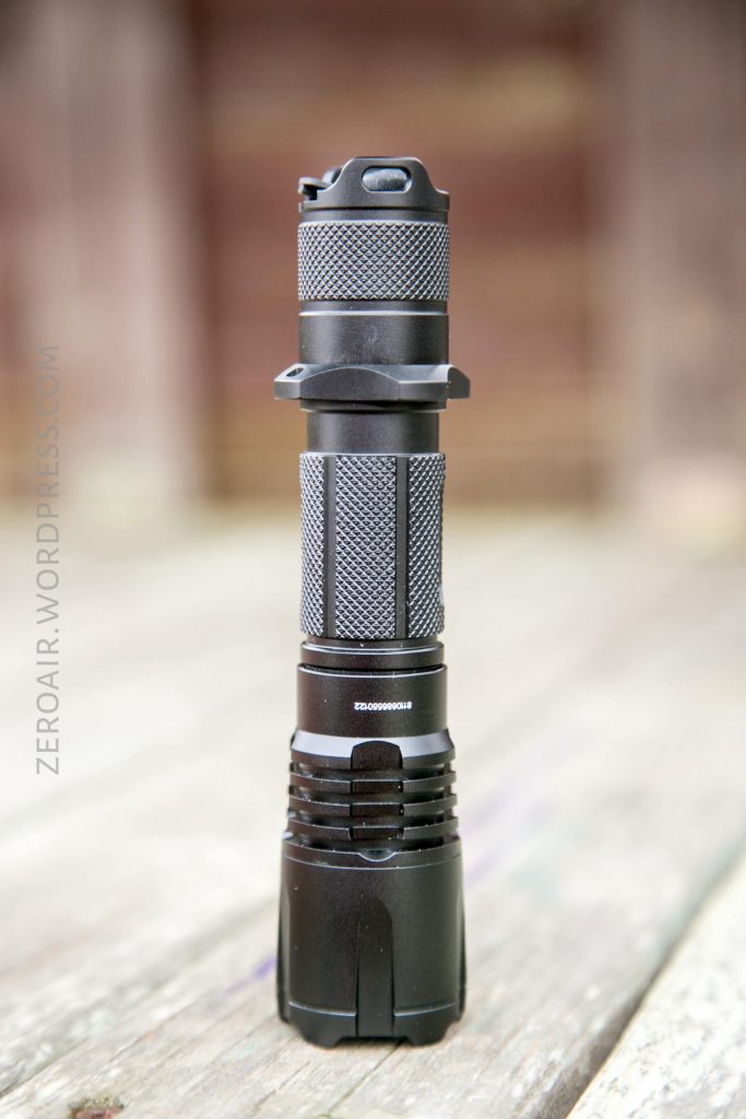 A black, textured tactical flashlight stands upright on a wooden surface, with a blurred brown background. The flashlight has a knurled grip and visible grooves for handling.