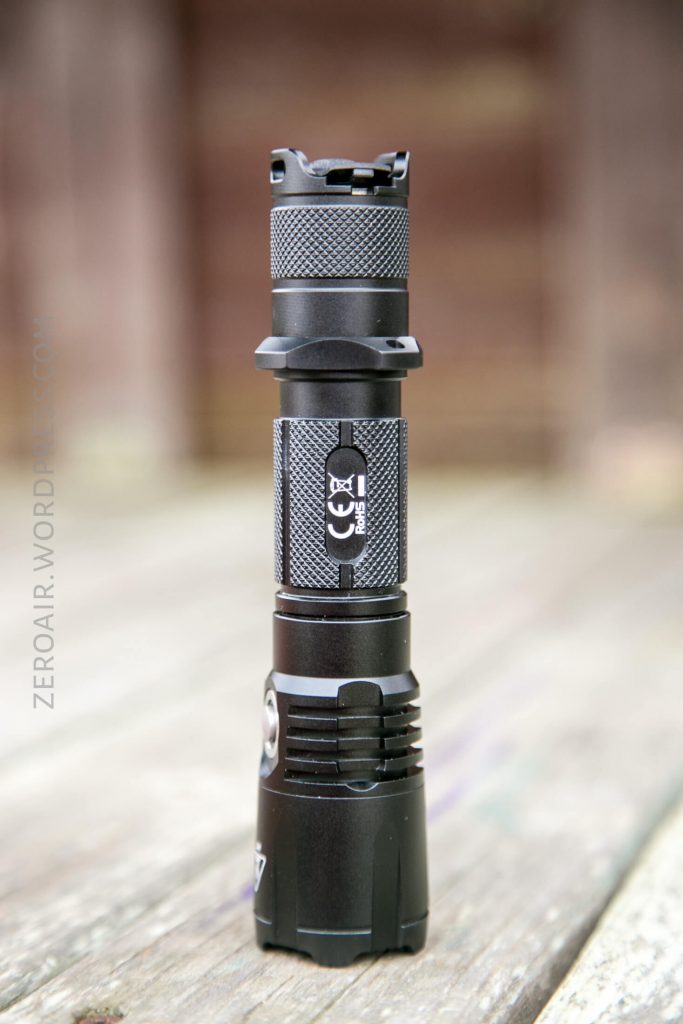 A black tactical flashlight stands upright on a wooden surface with a blurred brown background. The flashlight features a textured grip and a CE marking label on its body.