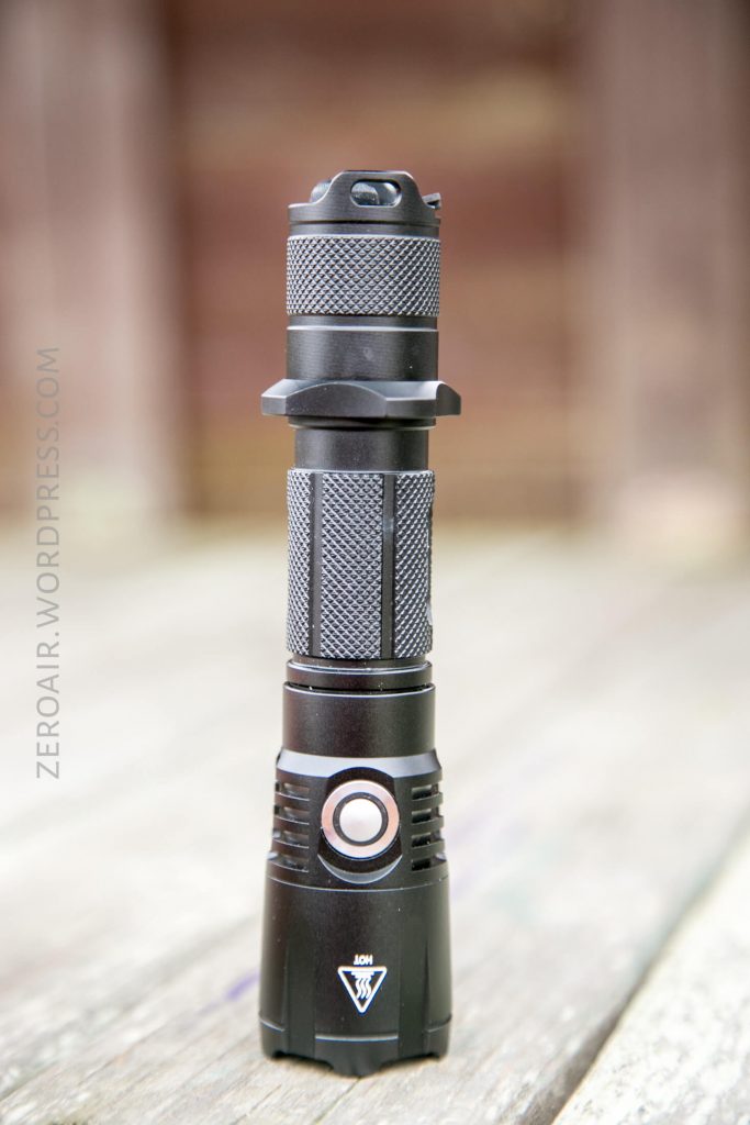 A black tactical flashlight with a textured grip and a button near the head is standing upright on a wooden surface. The background is blurred. The flashlight has a warning label for heat near the lens.