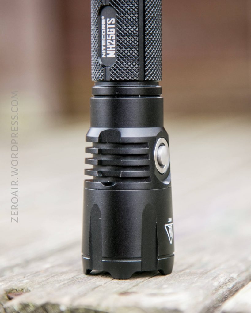 Close-up of a black Nitecore MH25GTS flashlight standing upright on a wooden surface, showing the textured grip, side button, and part of the manufacturers label.