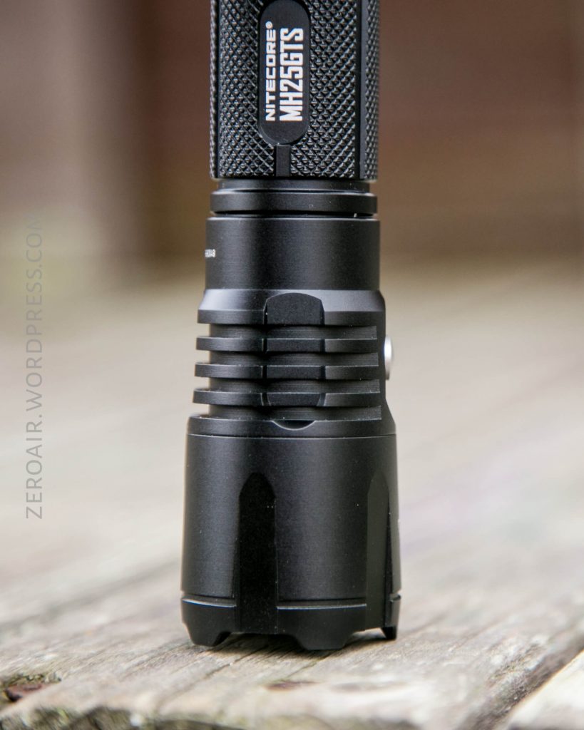 A close-up of a black Nitecore MH25S TS flashlight standing upright on a wooden surface, showing the textured grip and branding on the head of the flashlight.