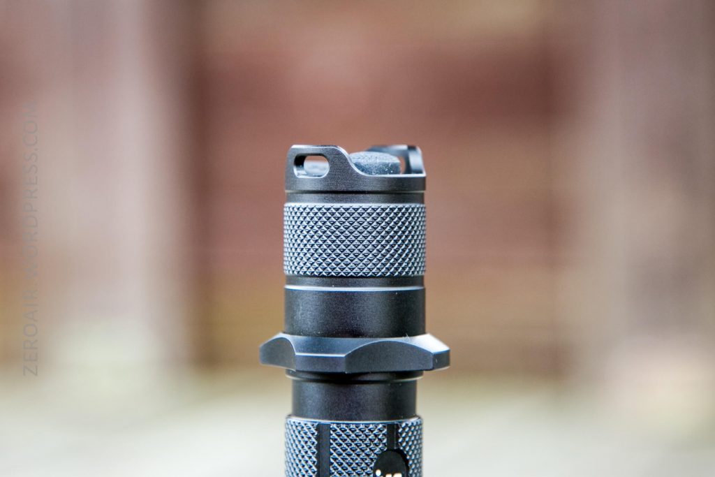 A close-up of the textured tailcap and body of a black flashlight, shown vertically against a blurred brown background. The image focuses on the knurled grip and flat base of the flashlight.