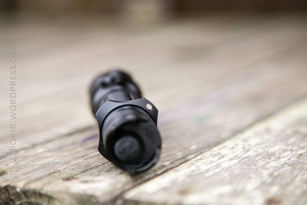A black flashlight lies on its side on a weathered wooden surface, photographed with the lens focused on the tailcap and the background blurred.