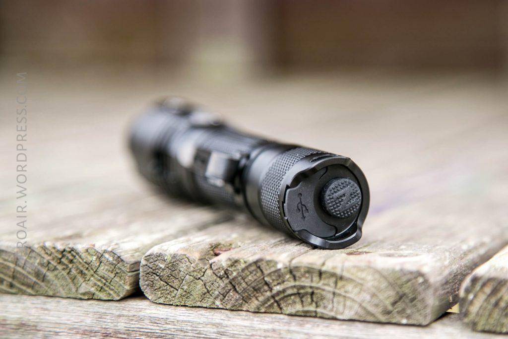 A black tactical flashlight lying on its side on a weathered wooden surface, with the focus on the textured tail switch and a visible USB port cover.