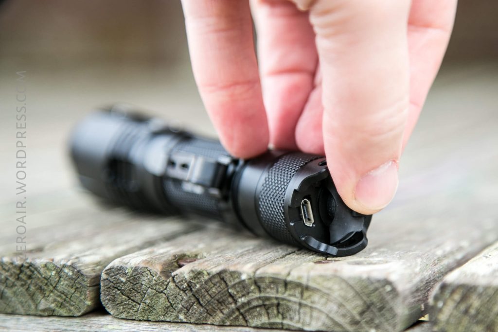 A hand lifts the rubber cover of a black flashlight to reveal a micro USB charging port. The flashlight rests on a weathered wooden surface.