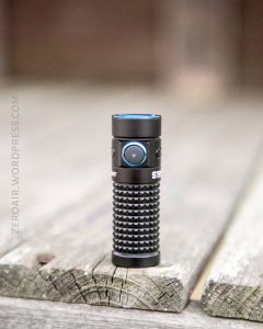 A black, compact flashlight with a textured grip and a blue button stands upright on a weathered wooden surface. The text ZEROAIR.WORDPRESS.COM is visible along the left side.