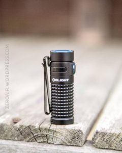 A small black Olight flashlight with a textured grip and side clip stands upright on a weathered wooden surface. The image background is blurred.
