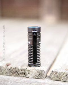 A small black flashlight with a textured grip stands upright on a weathered wooden surface. The flashlight has a black clip on its side. The background is blurred.
