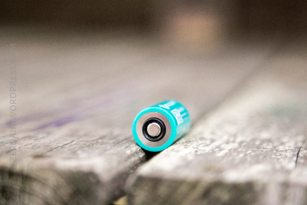 A teal cylindrical battery lying horizontally on a weathered wooden surface, with the positive terminal facing the camera. The background is blurred.