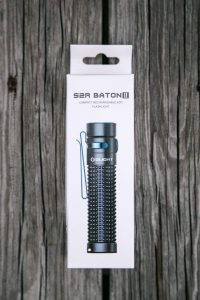 A white retail box for the Olight S2R Baton II compact rechargeable EDC flashlight rests on weathered wooden planks. The box displays an image of the black flashlight with a blue pocket clip.