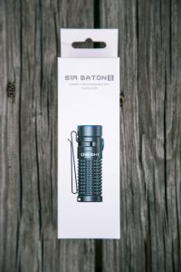 A white box with an image of a compact black flashlight labeled “Olight” on the front. The box reads “S1R Baton II Compact Rechargeable EDC Flashlight” and is placed on a weathered wooden surface.