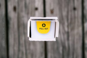 A close-up of an open white box with a yellow label inside that reads PULL OUT and a downward-pointing arrow. The background is a wooden surface with visible grain and knots.