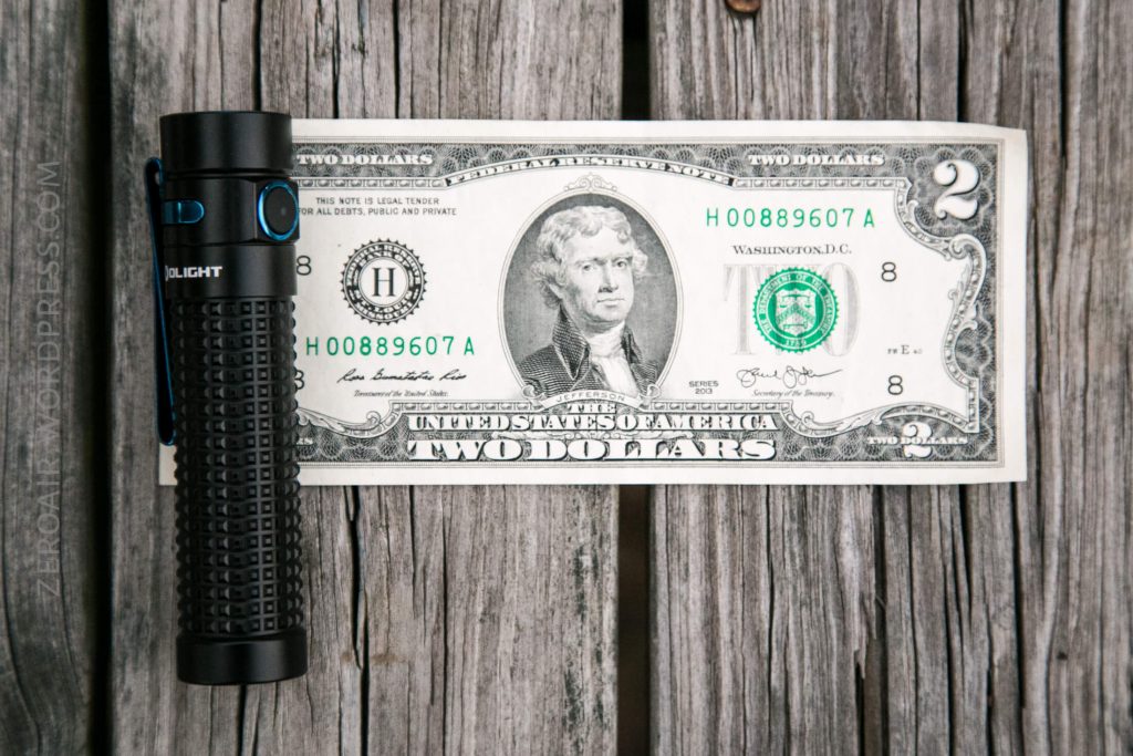 A black flashlight is placed vertically beside a two-dollar bill on a wooden surface. The bill displays Thomas Jefferson’s portrait and is positioned horizontally.